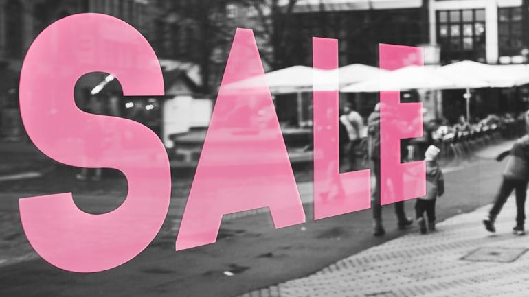 Das Bild zeigt im Hintergrund eine Einkaufszone. Es ist komplett überlegt mit einem pink-farbenen "Sale"-Schriftzug.